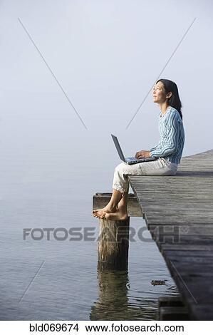 Picture - Asian woman holding laptop on edge of pier. Fotosearch