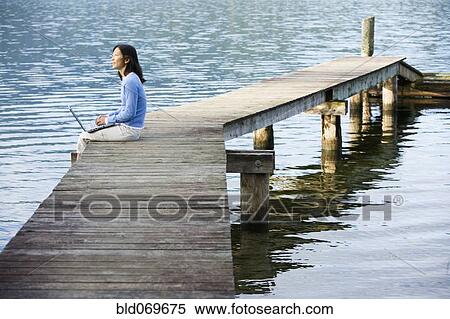 Stock Photography - Asian woman holding laptop on edge of pier. Fotosearch