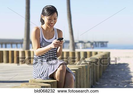 Stock Image - Asian woman sitting at beach using cell phone. Fotosearch