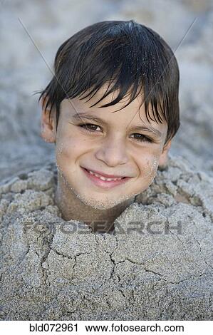 Stock Image - Close up of Hispanic boy buried in sand. Fotosearch
