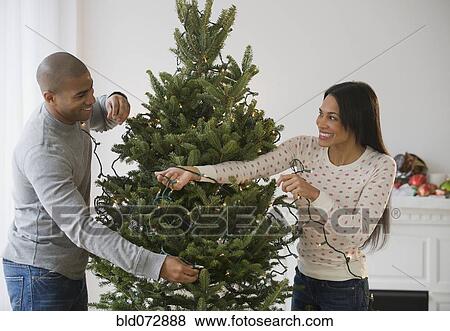 Stock Photo - Couple placing string lights on Christmas tree. Fotosearch