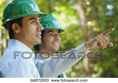 Stock Image - Hispanic businessmen in green hard hats pointing outdoors. Fotosearch