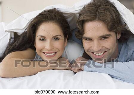 Stock Image - Hispanic couple smiling from under covers on bed. Fotosearch
