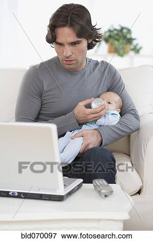 Stock Photo - Hispanic father feeding baby and looking at laptop . Fotosearch