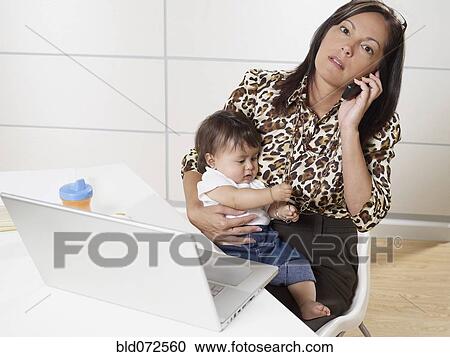 Stock Image - Hispanic working mother holding baby son and talking on phone. Fotosearch