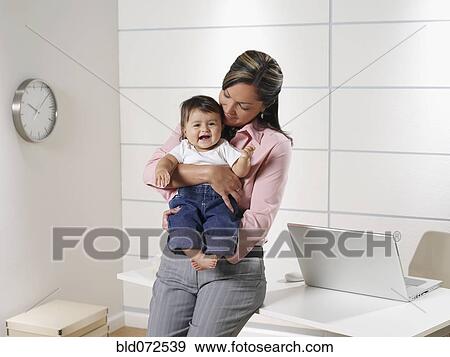 Stock Photo - Hispanic working mother holding baby son in home office. Fotosearch