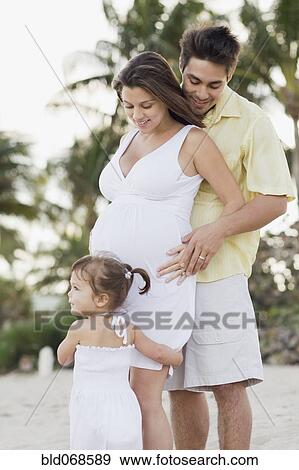 Stock Photo - Husband hugging pregnant wife and watching daughter. Fotosearch