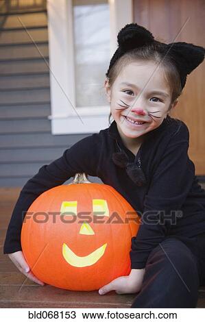 Stock Image - Mixed race young girl in cat costume holding Halloween pumpkin. Fotosearch