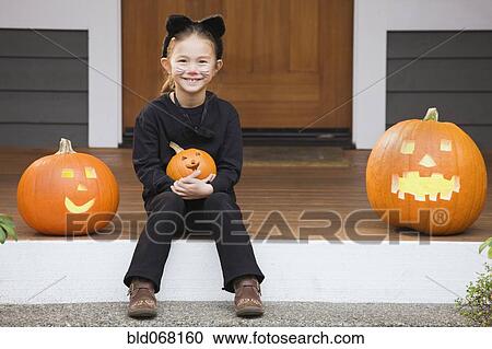 Stock Image - Mixed race young girl in cat costume holding Halloween pumpkin. Fotosearch