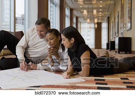 Stock Image - Multi-ethnic business people laying on floor reviewing blueprints. Fotosearch