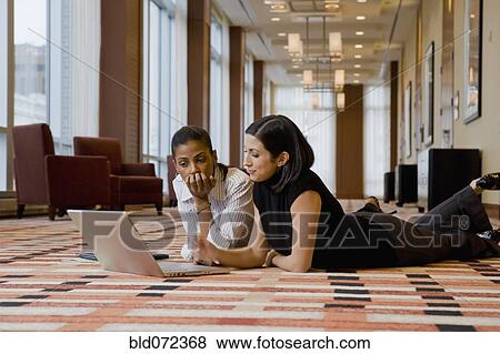 Stock Photo - Multi-ethnic businesswomen laying on floor working on laptop. Fotosearch