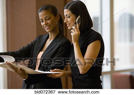 Stock Photo - Multi-ethnic businesswomen reviewing paperwork and talking on cell phone. Fotosearch