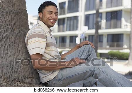 Young mixed race man sitting against tree View Large Photo Image Stock Photograph - Young mixed race man sitting against tree. Fotosearch