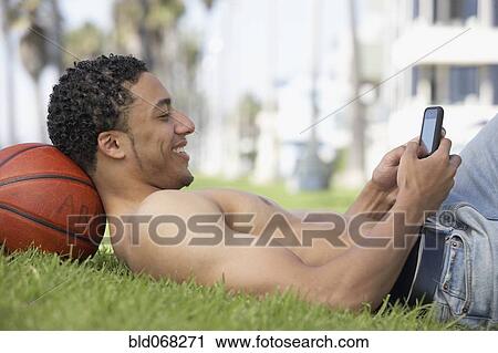 Stock Image - Young mixed race man with bare chest on grass with cell phone. Fotosearch