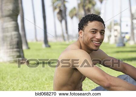 Stock Image - Young mixed race man with bare chest sitting on grass. Fotosearch