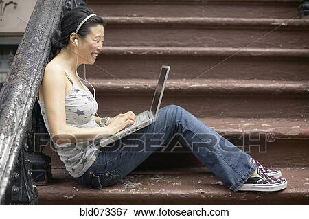 Stock Photo - Asian woman sitting on front steps with laptop. Fotosearch