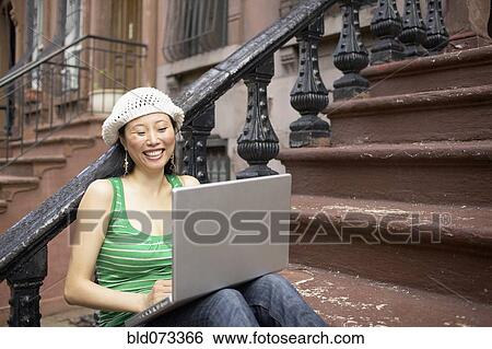 Stock Photograph - Asian woman sitting on front steps with laptop. Fotosearch