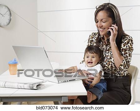 Picture - Hispanic working mother holding baby son and talking on phone. Fotosearch