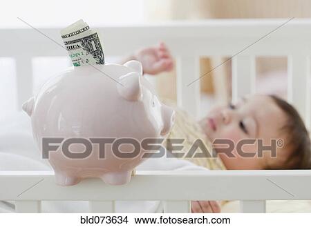 Picture - Mixed race baby girl laying in crib next to piggy bank. Fotosearch