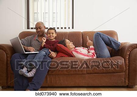 Stock Image - Antiguan man using laptop in living room with family. Fotosearch