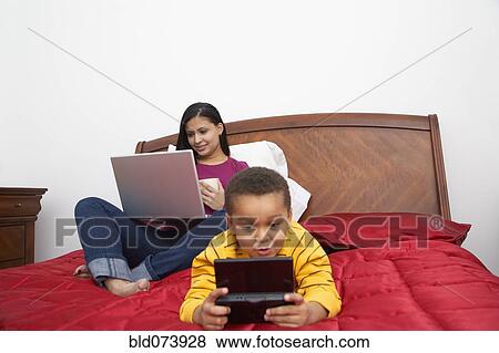 Stock Photo - "Dominican woman using laptop on bed, son playing video game". Fotosearch