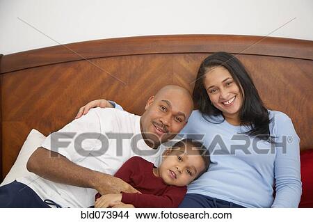 Picture - Mixed race boy lounging in bed with parents. Fotosearch