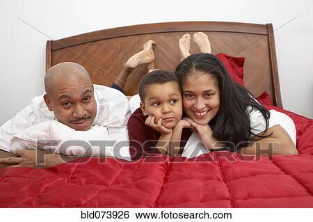 Stock Photograph - Mixed race boy lounging on bed with parents. Fotosearch