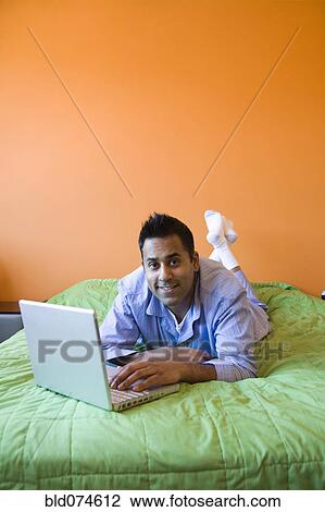 Stock Image - Mixed race man using laptop on bed. Fotosearch