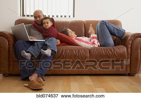 Stock Photo - Antiguan man using laptop in living room with family. Fotosearch