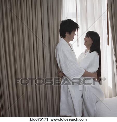 Stock Image - Asian couple in bathrobes hugging in hotel room. Fotosearch