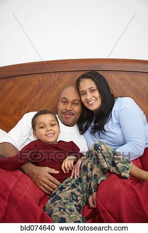 Stock Image - Mixed race boy lounging in bed with parents. Fotosearch