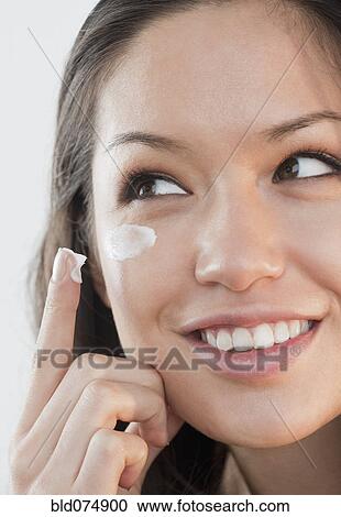 Stock Image - Mixed race woman applying cream under eyes. Fotosearch