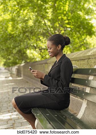 Stock Image - African businesswoman looking at cell phone on bench. Fotosearch