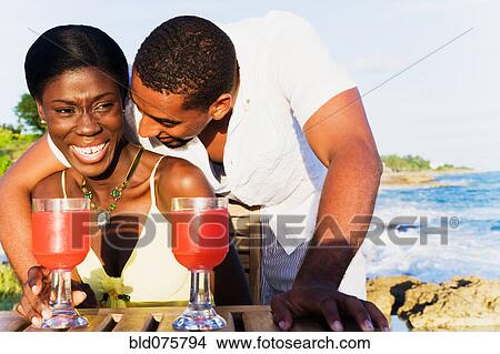 Picture - African couple drinking cocktails on patio overlooking ocean. Fotosearch