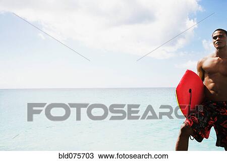 Stock Image - African man holding body board on beach. Fotosearch
