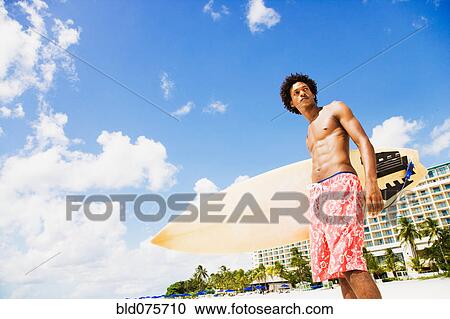 Stock Image - African man holding surfboard on beach. Fotosearch