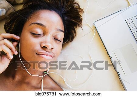 Stock Image - African woman listening to music on headphones connected to laptop. Fotosearch