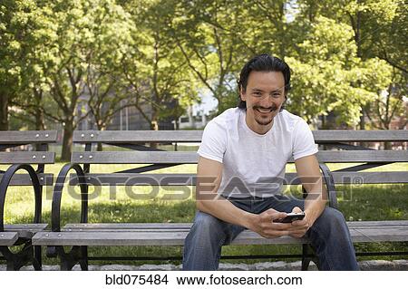 Picture - Mixed race man holding cell phone on park bench. Fotosearch