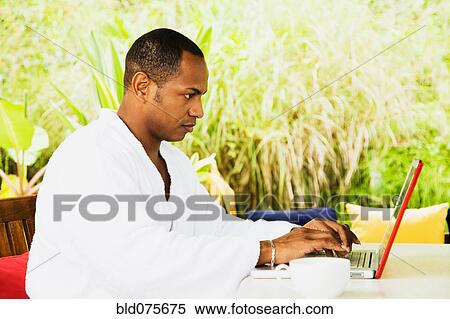 Stock Photography - Mixed race man using laptop in bathrobe on patio. Fotosearch