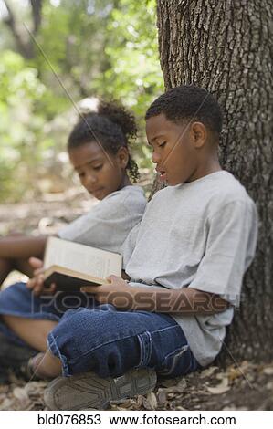 Stock Image - African boy and girl reading book against tree. Fotosearch