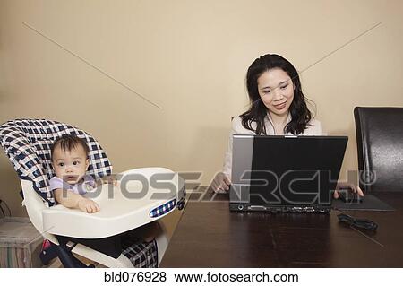 Chinese mother working on laptop next to baby in high chair View Large Photo Image Stock Photo - Chinese mother working on laptop next to baby in high chair. Fotosearch