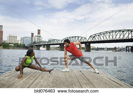 Stock Image - Couple stretching on urban boardwalk along river. Fotosearch
