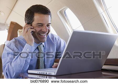 Stock Image - Hispanic businessman using laptop and cell phone on airplane. Fotosearch