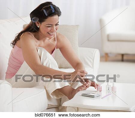 Stock Image - Hispanic woman painting toenails on sofa. Fotosearch