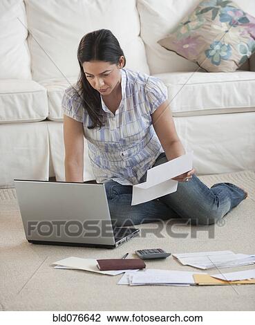 Stock Image - Hispanic woman paying bills with laptop on living room floor. Fotosearch