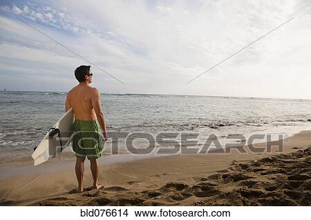 Picture - Japanese man holding surfboard on beach. Fotosearch