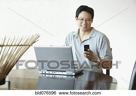 Japanese man using laptop and checking cell phone at table View Large Photo Image Stock Photograph - Japanese man using laptop and checking cell phone at table. Fotosearch