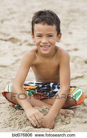 Stock Photograph - Mixed race boy in swim trunks sitting on beach. Fotosearch