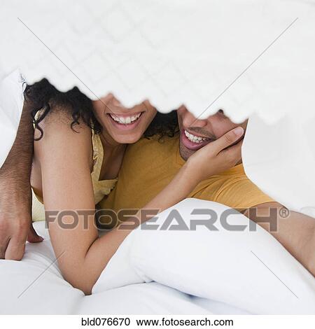 Stock Image - Smiling couple hiding under bed sheet. Fotosearch