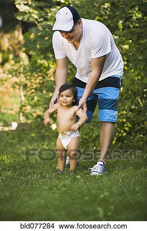 Picture - Hispanic father helping baby son to walk in grass. Fotosearch